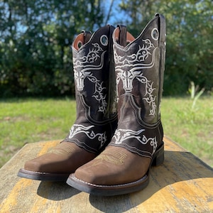 May include: A pair of brown and black cowboy boots with white embroidered bull and scroll designs. The boots have square toes and are displayed on a wooden surface, with a blurred green background.