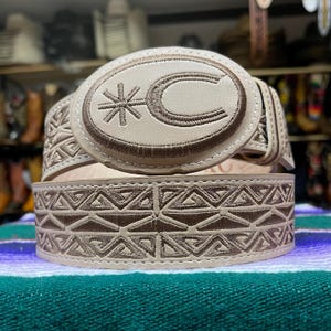 May include: A beige leather belt with a decorative buckle. The buckle features a stitched design with a star and the letter "C". The belt has a geometric pattern stitched along its length. The belt is displayed on a striped surface.