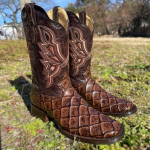 Puede incluir: Un par de botas de vaquero de cuero marrón con costuras intrincadas y un patrón texturizado en la parte inferior. Las botas tienen una punta puntiaguda y un tacón apilado, adecuado para el uso occidental. Las botas se exhiben al aire libre sobre una superficie cubierta de hierba.