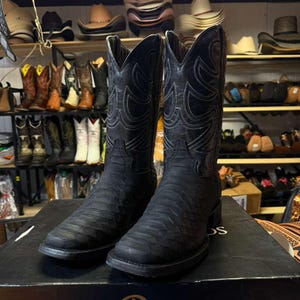 May include: A pair of black cowboy boots featuring a textured, scale-like pattern on the foot and a smooth leather shaft with intricate white stitching. The boots have a square toe design and are positioned on a black box in a retail setting.