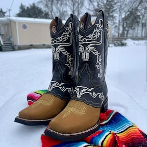May include: Black and tan cowboy boots with white bull embroidery. The boots have a square toe and are displayed on a colourful serape blanket in a snowy outdoor scene.