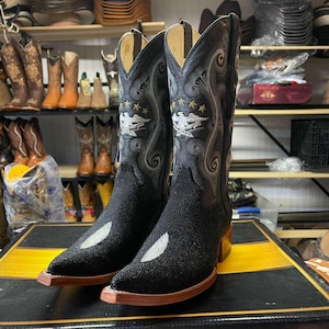 May include: A pair of black cowboy boots with detailed grey and gold embroidery. The boots have a textured toe and heel, with an eagle and star design. The boots are displayed on a black and yellow surface.