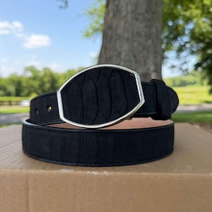 May include: A black belt featuring a textured surface with a repeating scale-like pattern and a large, smooth, silver-toned metal buckle shaped like an elongated hexagon. The belt strap is made of a matte black material with visible stitching.