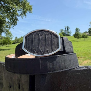 May include: A black belt featuring a textured, scale-like pattern throughout the strap and a large, oval-shaped buckle with a silver-toned metal frame surrounding a matching black, textured inlay that mimics the appearance of reptile skin.