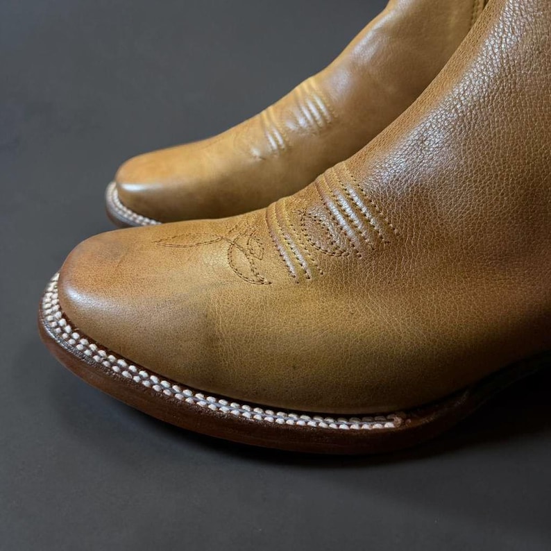 May include: Close-up of a pair of brown leather cowboy boots. The boots have a rounded toe, decorative stitching, and a white stitched welt. The leather has a worn, textured appearance.