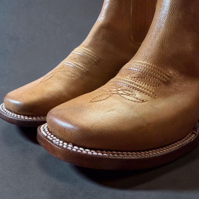 May include: Close-up of a pair of tan leather cowboy boots. The boots have a square toe design with decorative stitching on the vamp and shaft. The soles are brown with white stitching around the edges. The leather has a worn, textured appearance.