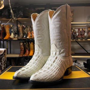 Könnte beinhalten: Ein Paar cremefarbene Cowboy-Stiefel. Der Fuß jedes Stiefels hat ein strukturiertes, krokodilartiges Muster, während der Schaft ein gesticktes Design aufweist. Sie haben schwarze Sohlen und Absätze und werden auf einer schwarz-gelben Oberfläche präsentiert.