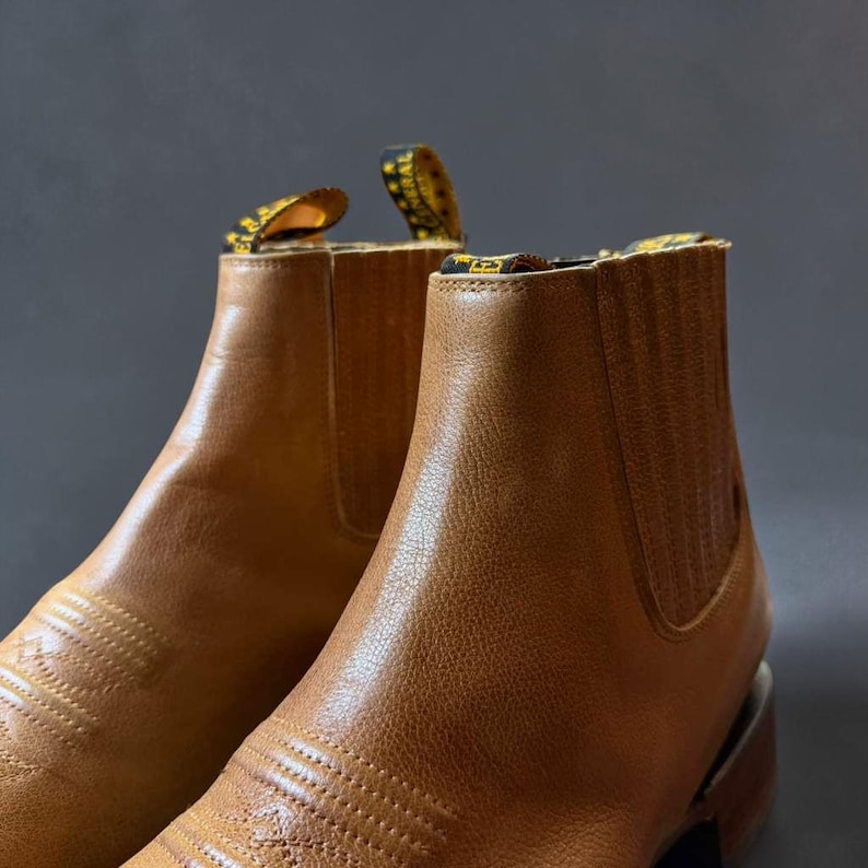 May include: Close-up of a pair of tan leather ankle boots. The boots have a classic Chelsea boot design with elastic side panels and pull tabs. The leather has a textured appearance, and the boots are set against a gray background.