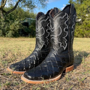 May include: A pair of black cowboy boots with a square toe and a textured pattern on the vamp. The boots have white stitching details and a brown sole. The boots are standing on grass.