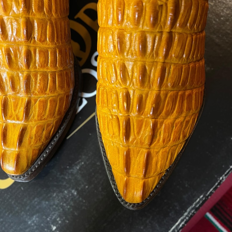 May include: Close-up of two yellow cowboy boots with a textured, crocodile-like pattern. The boots have pointed toes and dark brown trim. The leather has a glossy finish, and the boots appear to be new.