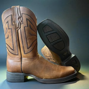 May include: A pair of brown leather cowboy boots with decorative stitching and a square toe. One boot is upright, while the other is angled to show the black sole. The boots have a classic Western design.