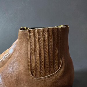 May include: Close-up of a brown leather Chelsea boot. The boot features a textured, pleated design on the side panel and a contrasting yellow and black patterned trim at the top. The boot is set against a dark gray background.