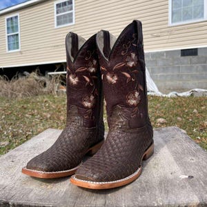 May include: A pair of brown leather cowboy boots. The foot features a woven pattern, while the shaft has a floral embroidered design. The boots have a square toe and a light brown sole.