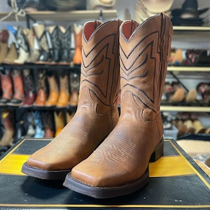 May include: A pair of brown leather cowboy boots with square toes and decorative stitching. The boots have a worn, distressed appearance and are displayed on a black and yellow striped surface. The background shows a variety of other boots.