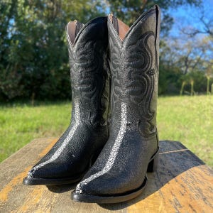 May include: A pair of black cowboy boots with a pointed toe and decorative stitching. The boots feature a white stripe down the front and are made of a textured material. The boots are displayed on a wooden surface.