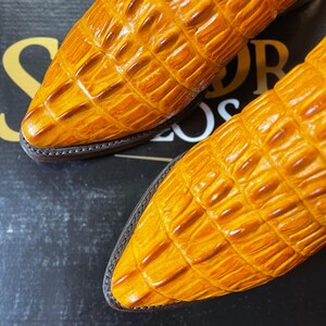 May include: Close-up of two yellow-orange leather shoes with a textured, crocodile-like pattern. The shoes have pointed toes and brown soles with a stitched border. The image is a close-up, focusing on the texture and color of the footwear.