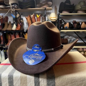 Könnte beinhalten: Ein brauner Cowboyhut mit breiter Krempe und dekorativem Band. Der Hut hat ein Etikett mit der Aufschrift "Katanna Gold Line". Der Hut wird auf einer gemusterten Decke präsentiert, mit Regalen voller Cowboy-Stiefel und -Hüte im Hintergrund.
