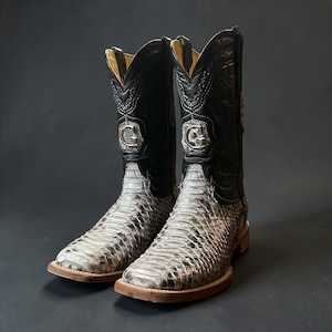 May include: A pair of cowboy boots with a snakeskin pattern on the lower part and black leather uppers. The boots have a silver emblem and a light brown sole. The background is a solid dark gray.