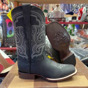 May include: Black leather cowboy boots with detailed white embroidery. The boots feature a square toe and a brown sole. The heel is also brown. The boots are displayed on a wooden surface. A classic Western style.