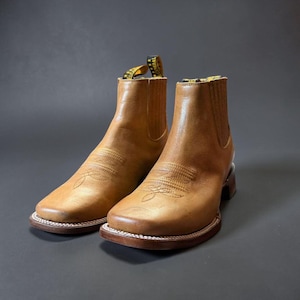 May include: A pair of tan leather ankle boots with square toes and decorative stitching. The boots have a pull tab with yellow and black text. The soles are brown with a white stitched border. The boots are set against a dark gray background.