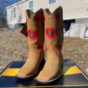 May include: Tan cowboy boots featuring red sunflower embroidery. These boots have a square toe, a stacked heel, and white stitching. They are made from a suede-like material. The boots are approximately 30 cm tall.