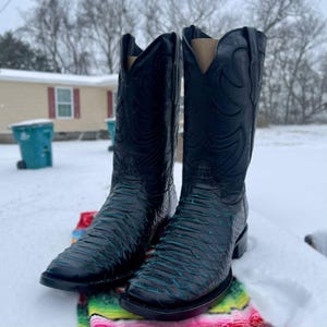 May include: A pair of black cowboy boots with a textured design on the foot and shaft. The boots are resting on a colourful blanket in a snowy outdoor setting. The boots have a square toe and a tall shaft.