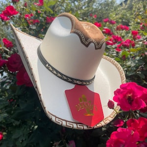 Könnte beinhalten: Ein cremefarbener Cowboyhut mit braunem Wildleder-Oberteil und Zierleisten. Der Hut hat ein schwarzes Band mit goldenen Akzenten und ein rotes Etikett mit goldener Schrift. Umgeben von roten Rosen.