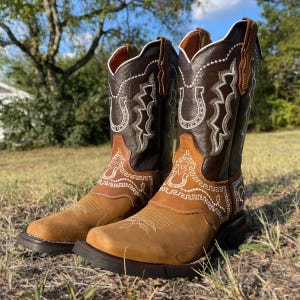 May include: A pair of brown and tan cowboy boots with decorative white stitching. The boots have a square toe, a stacked heel, and a horseshoe design. The boots are made of leather and are displayed outdoors.
