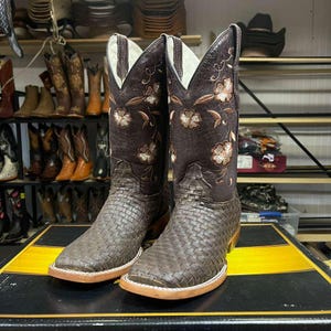 May include: A pair of brown leather cowboy boots with floral embroidery and a woven pattern on the foot. The boots have a square toe and a light-coloured sole. The shaft features embroidered flowers.