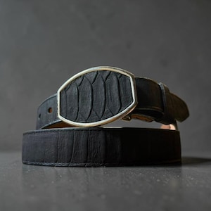 May include: A black belt featuring a textured surface with a repeating scale-like pattern. The belt includes a prominent oval-shaped buckle with a metallic silver-toned rim that frames a matching black textured inlay, creating a cohesive look.