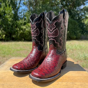 Peut inclure: Une paire de bottes de cowboy rouges et noires. Les bottes ont un pied en cuir brun-rouge texturé et une tige en cuir noir avec des coutures décoratives. Les bottes sont exposées sur une surface en bois.