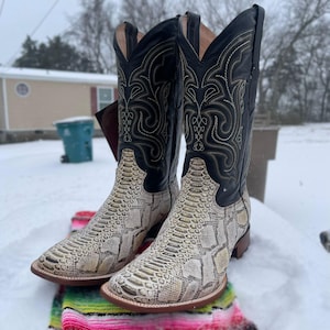 May include: Cowboy boots featuring a snakeskin design on the foot and a black leather upper with decorative stitching. The boots are placed on a colourful blanket in a snowy outdoor setting.