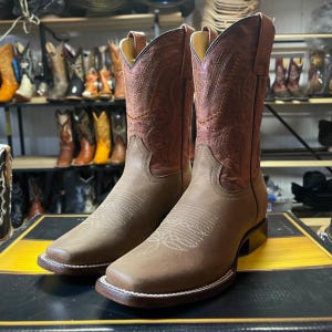 May include: A pair of brown leather cowboy boots with a square toe and decorative stitching. The boots have a reddish-brown upper with intricate embossed patterns and a light brown lower section. The boots are displayed on a black and yellow surface.
