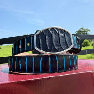 May include: A black belt featuring a textured, scale-like pattern with vertical bright blue stripes. The belt includes a large, oval-shaped silver-toned metal buckle with a smooth, polished frame that secures the strap in place.