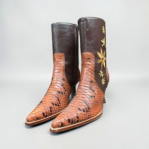 Könnte beinhalten: Ein Paar braune Lederstiefel. Der untere Teil der Stiefel hat ein Schlangenmuster, während der obere Teil einfarbig braun ist. Sie haben spitze Zehen und kleine Absätze. Ein Stiefel hat gestickte gelbe Blumenmuster.
