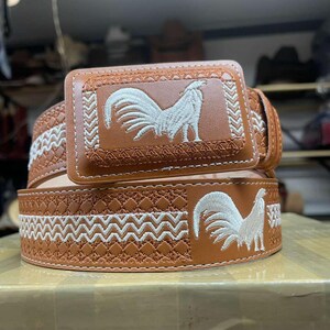 Könnte beinhalten: Ein brauner Ledergürtel mit rechteckiger Schnalle. Der Gürtel und die Schnalle weisen weiß gestickte Hahn-Designs auf. Der Gürtel hat ein dekoratives Nahtmuster entlang der Kanten. Die Schnalle hat einen helleren Braunton als der Gürtel.