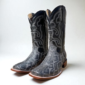 May include: A pair of black and grey cowboy boots with a snakeskin pattern on the foot and lower leg. The boots have decorative stitching and a square toe. The soles are brown with a red edge.