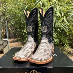 May include: A pair of cowboy boots with a snakeskin pattern on the foot and black leather uppers. The boots have a square toe and a decorative silver emblem. The soles are a light brown color.