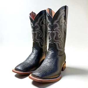 May include: A pair of black cowboy boots with a textured pattern on the foot and shaft. The boots have white stitching and a square toe. The soles are brown with a light-coloured welt. The interior is a light pink colour.
