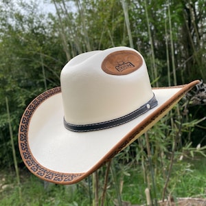 Könnte beinhalten: Ein cremefarbener Cowboyhut mit einem braunen Lederband und Krempenbesatz. Der Hut hat ein schwarzes Lederband um die Krone und einen braunen Lederaufnäher mit dem Wort "Rodeo".