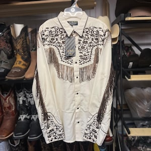 Puede incluir: Camisa vaquera color crema con bordados florales marrones y detalles de flecos. La camisa de manga larga presenta una parte delantera abotonada y un cuello. Una etiqueta cuelga de la camisa. La camisa se exhibe en una percha.