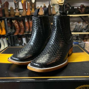 May include: Black leather ankle boots with a woven design. The boots feature a square toe, a light brown sole, and a small heel. They are presented on a black box with a yellow stripe.