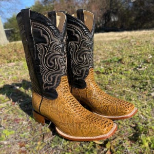 May include: A pair of cowboy boots with a textured, tan-coloured foot and a black leather upper. The black upper features intricate white and grey stitching. The boots have a square toe and a brown heel.