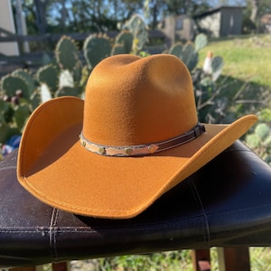 May include: A tan cowboy hat with a wide brim and a rounded crown. The hat has a brown leather band with decorative studs. The hat is made of a felt-like material. The hat is designed for sun protection and style.