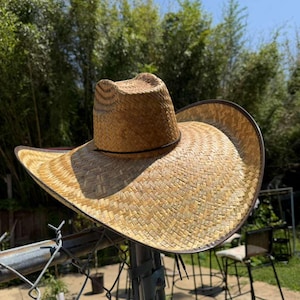 Op de afbeelding: Een beige strooien cowboyhoed met een ingedeukte kroon en een brede, gebogen rand. De hoed is voorzien van een donkerbruine bies langs de rand van de hoed en een dunne, donkerbruine band rond de basis van de kroon voor een gestructureerde look.