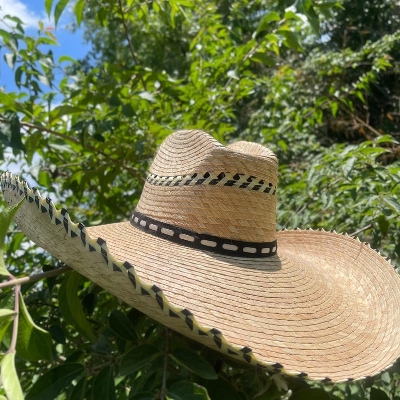 May include: A wide-brimmed straw hat with a light brown hue. The hat features a black and white band around the crown and decorative stitching along the brim. The hat is set against a backdrop of green foliage and a blue sky.