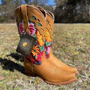 May include: Tan leather cowboy boots with vibrant floral embroidery. The boots feature a square toe and decorative stitching. A tag with the text "CENTENARIO" is attached to one boot.