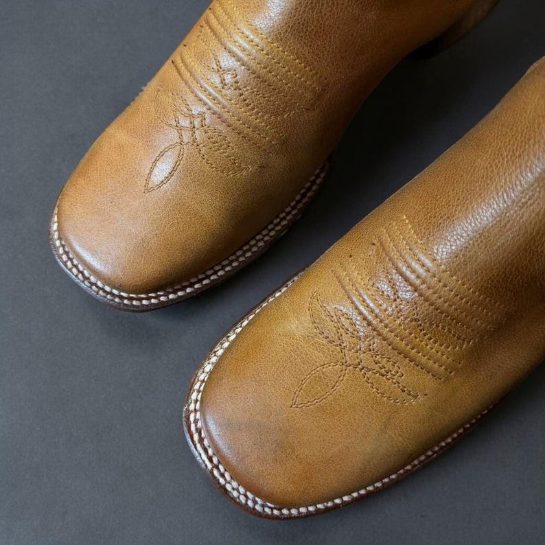 May include: Close-up of a pair of brown leather cowboy boots. The boots have a square toe design with decorative stitching on the vamp and shaft. The soles are stitched with white thread, and the leather has a worn appearance.