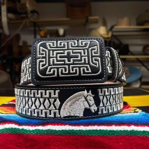 May include: Black leather belt with intricate white embroidery. The belt buckle features a geometric design, while the belt itself showcases a horse head and geometric patterns. A stylish accessory for any outfit.