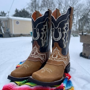 May include: A pair of brown, black, and tan cowboy boots with white decorative stitching and a horseshoe design. The boots have a square toe and are set on a colourful blanket in a snowy outdoor setting.